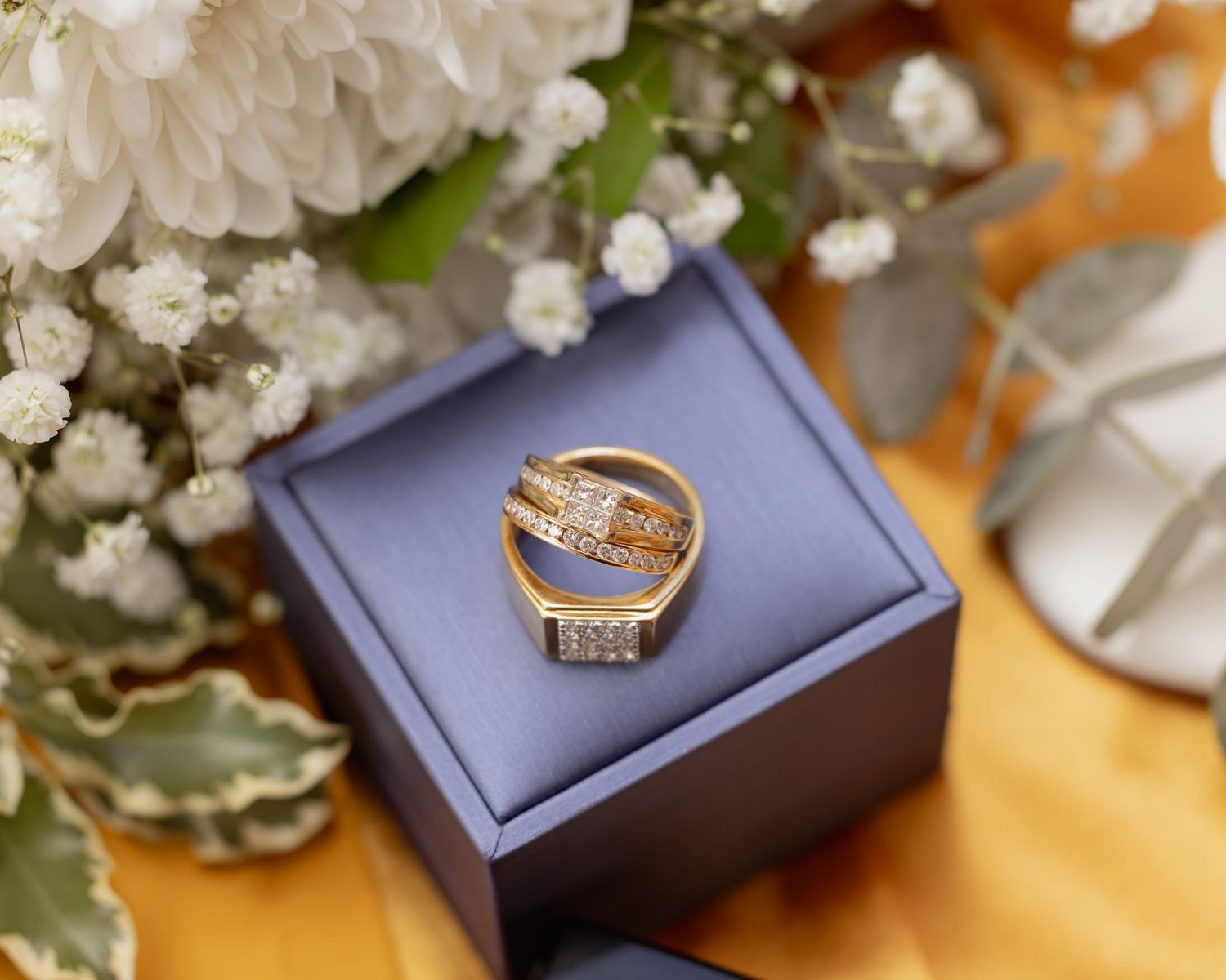 Gold wedding ring in navy blue box with white flowers and baby's breath displayed on orange fabric background.