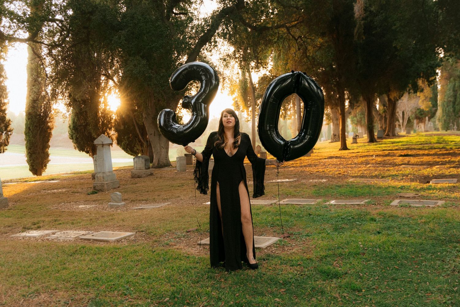 Black balloons in the shape of numbers 30 float in a sunlit park during golden hour with trees in the background.