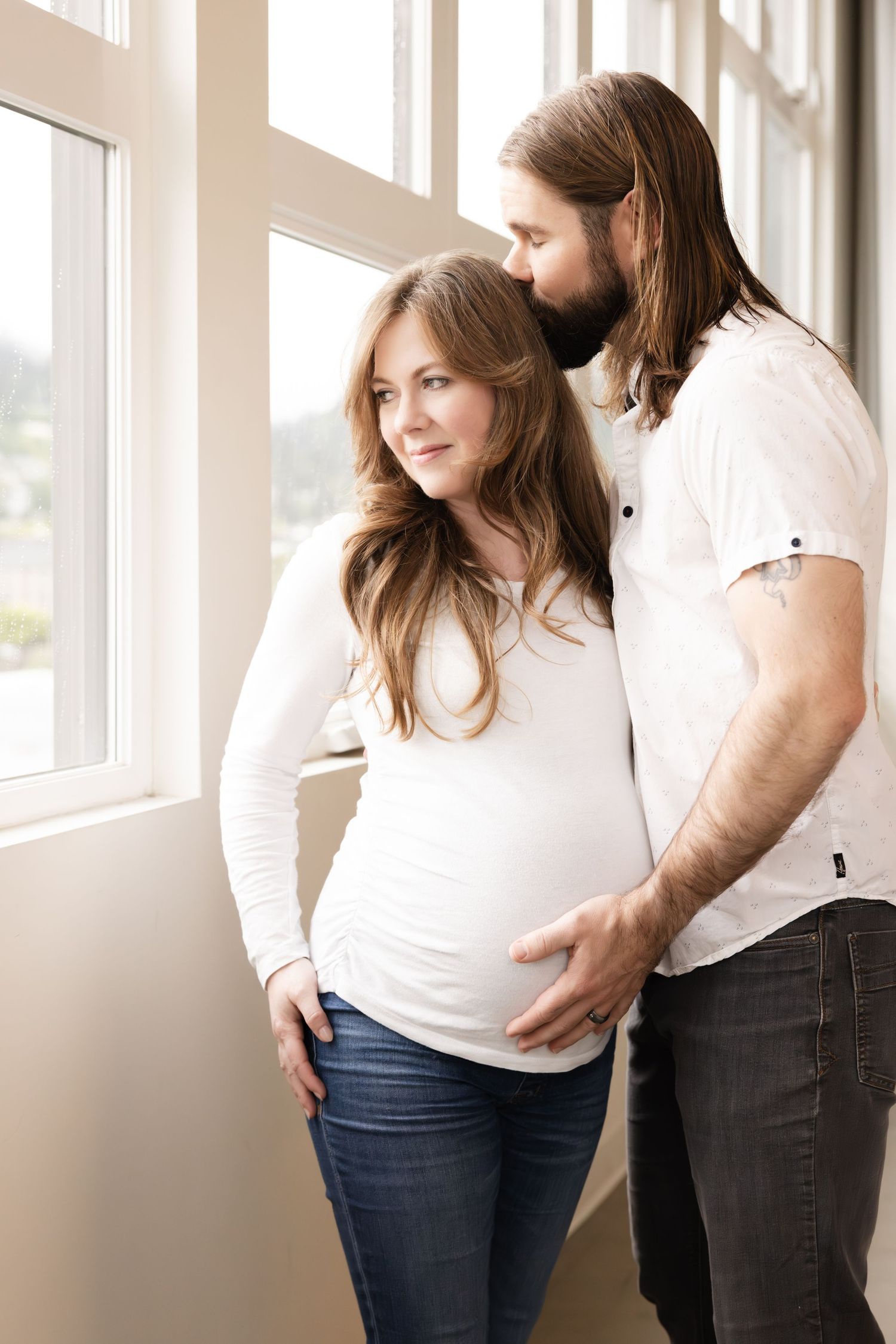 A couple stands by a bright window as sunlight illuminates their tender moment during a maternity photoshoot.