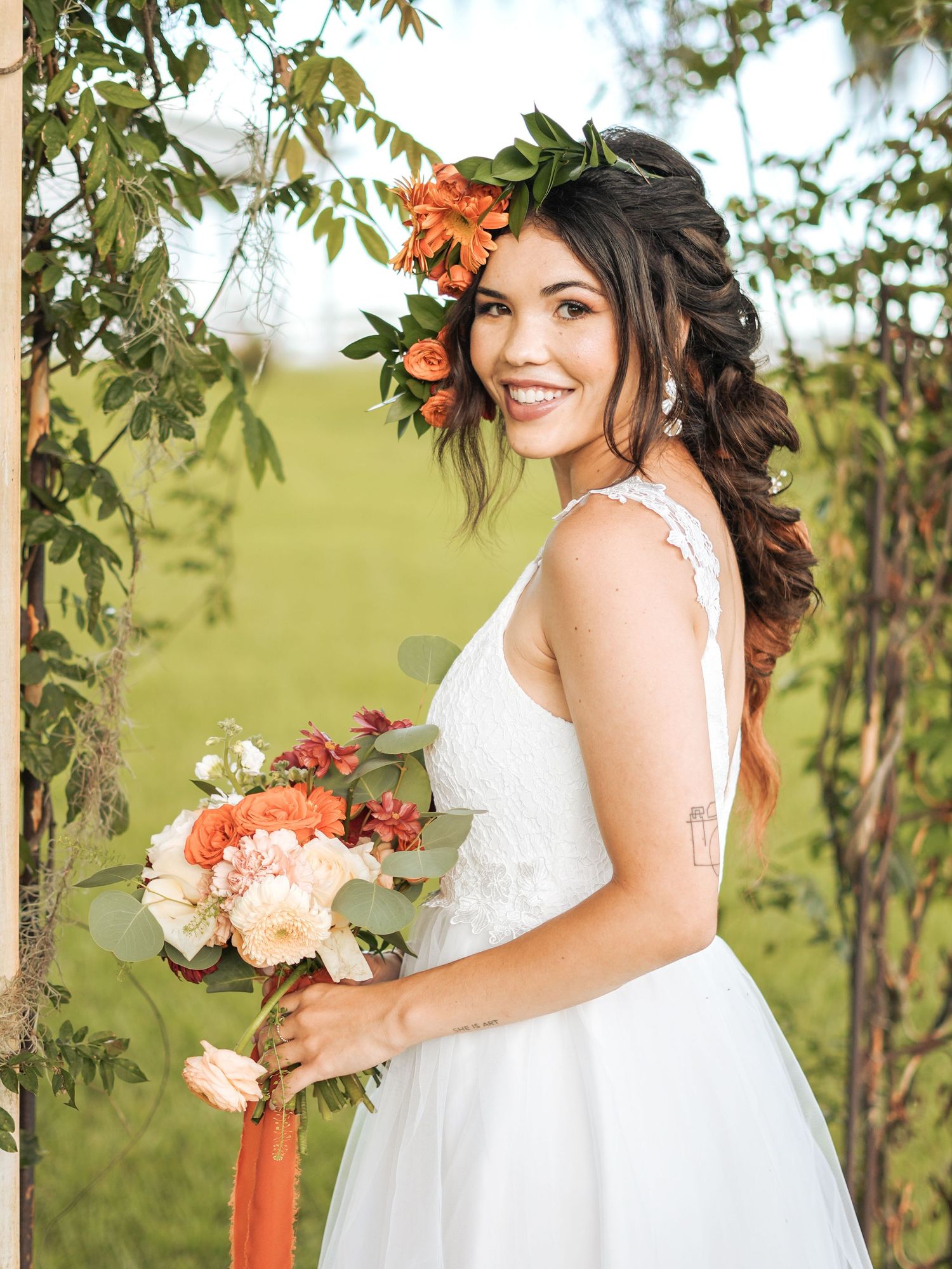 A bride in a white wedding dress holds a coral and peach bouquet while posing under green foliage outdoors.