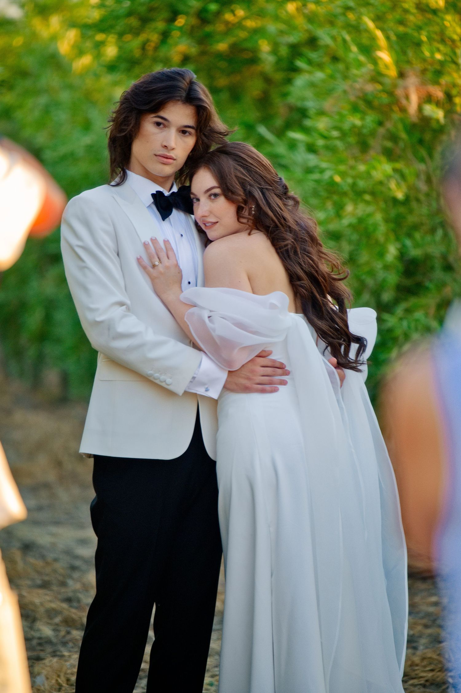 A romantic couple in formal attire embraces outdoors, with the bride wearing a flowing white dress against a natural backdrop.