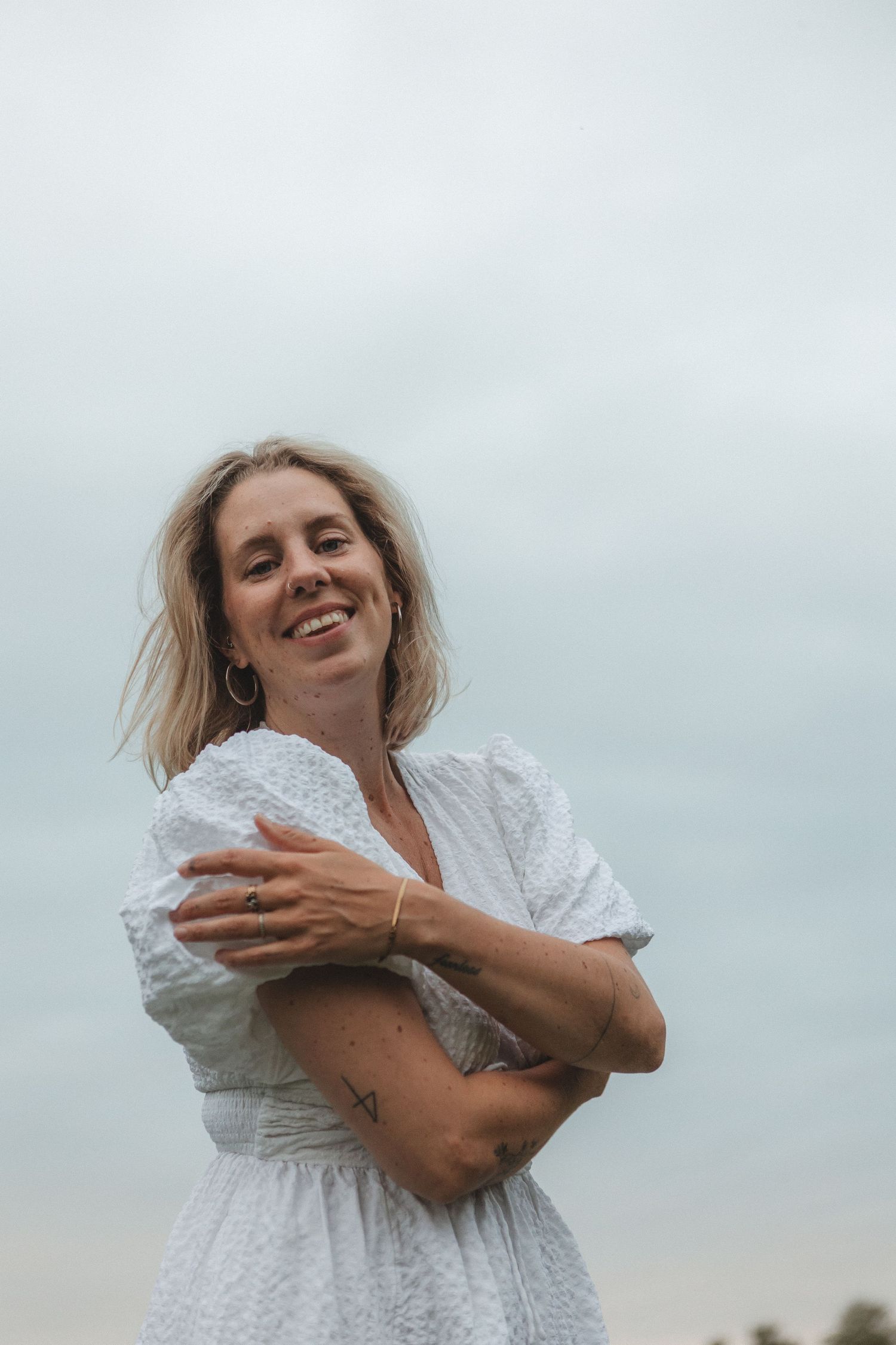 Person wearing a white eyelet dress laughing against a cloudy sky background.