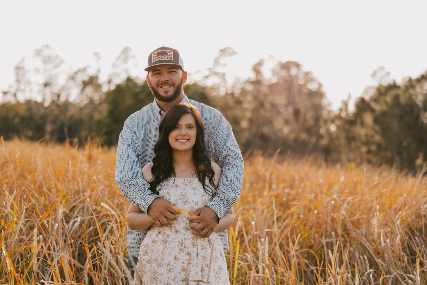 Couple embraces in golden wheat field during sunset with trees in background at Bartow Peace River park