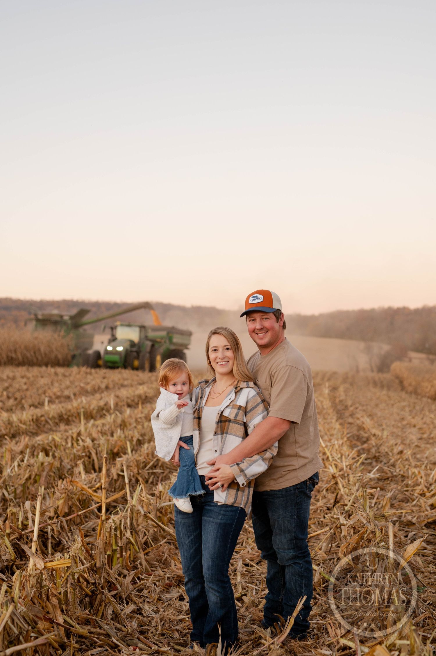 A family poses together in a harvested corn field at sunset with farm equipment in background.