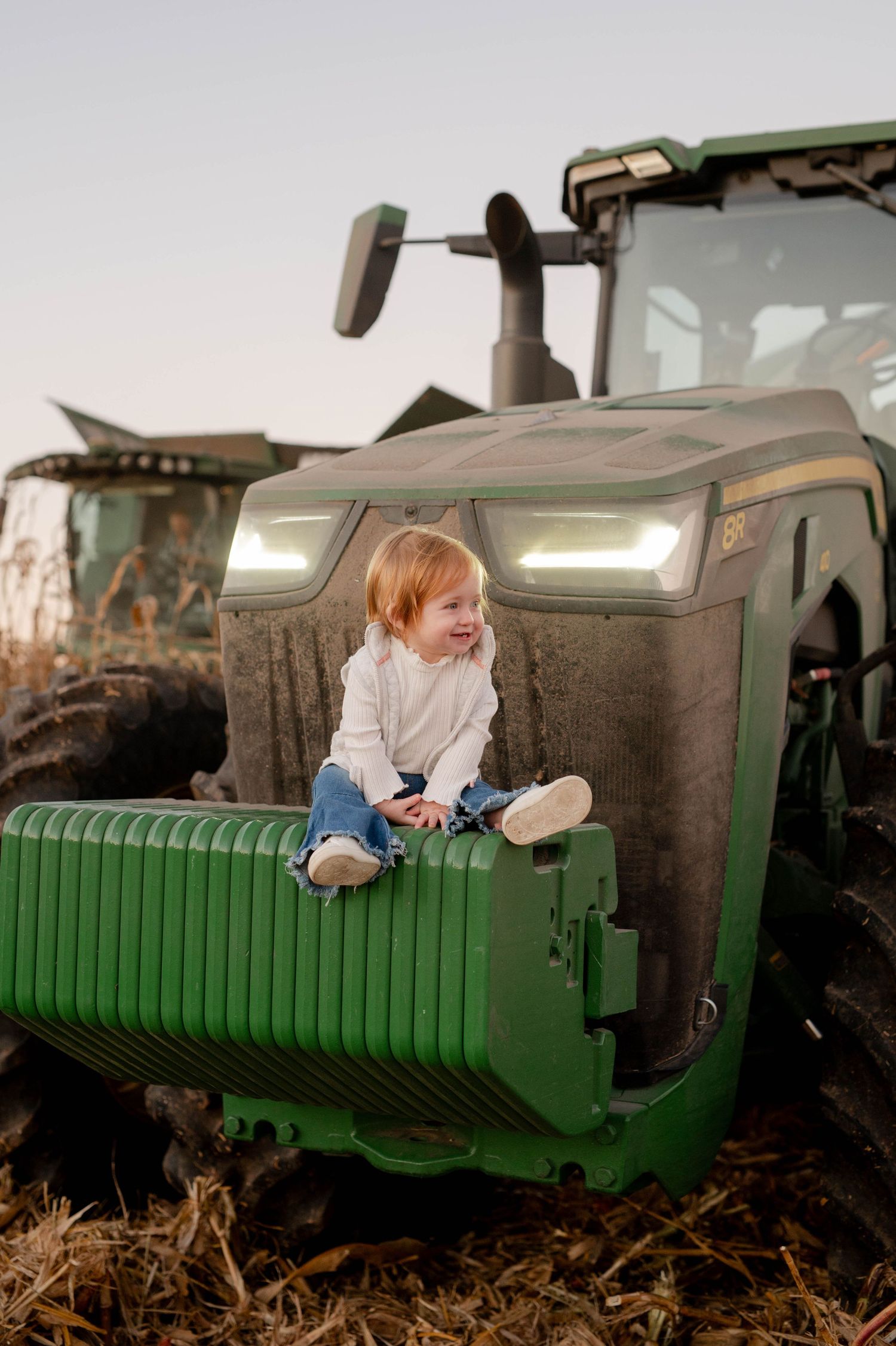 Smiling child perched on the front of a large green tractor in a cornfield.