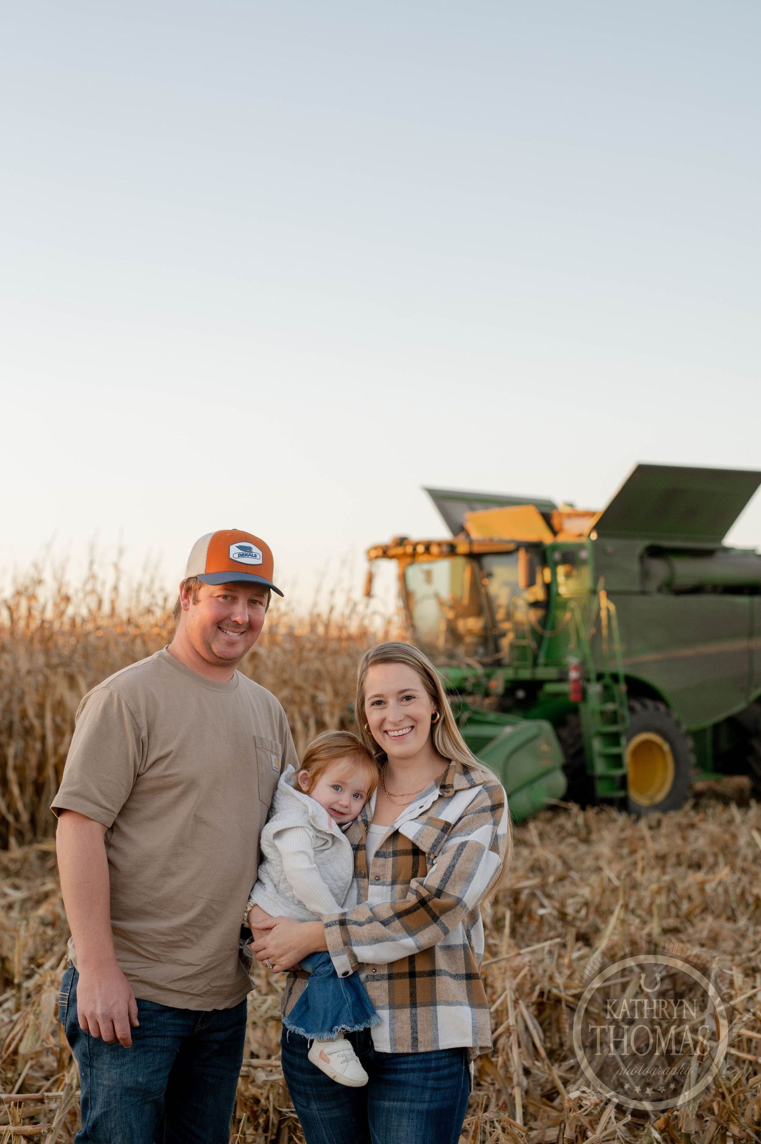 Family poses together by combine harvester with cornfield backdrop at dusk.