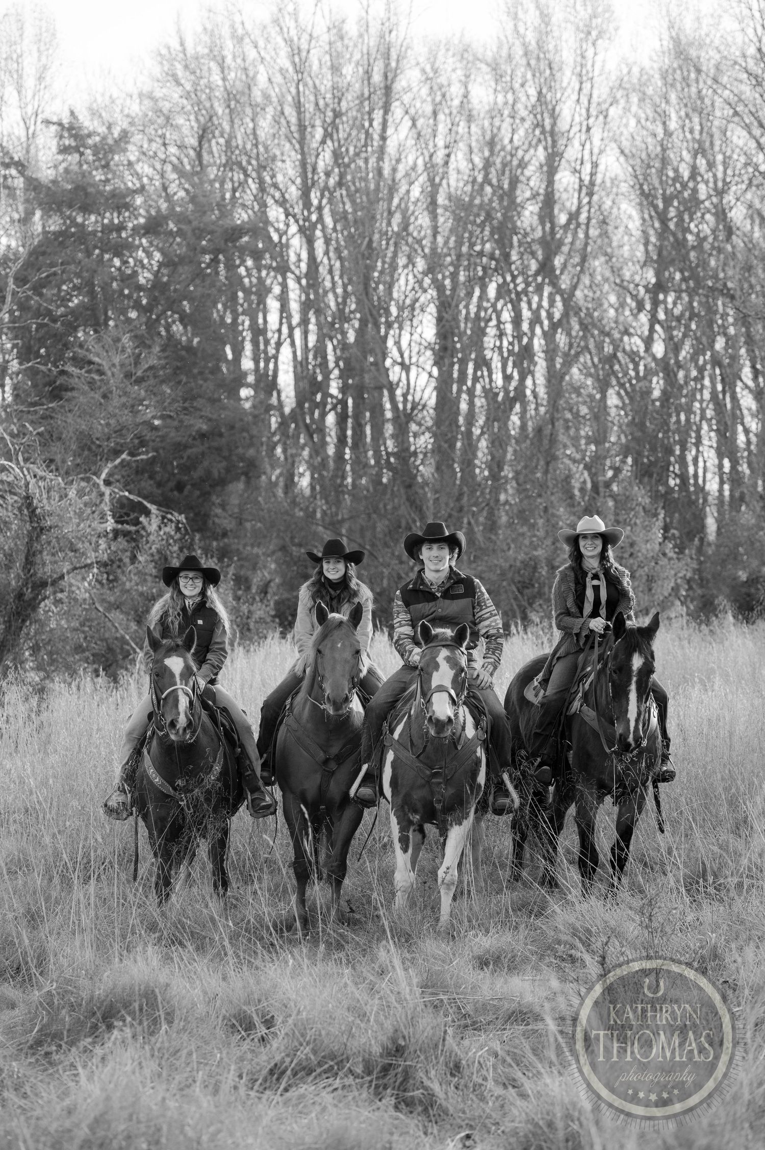 Black and white photo of four riders on horseback in a line against bare trees.