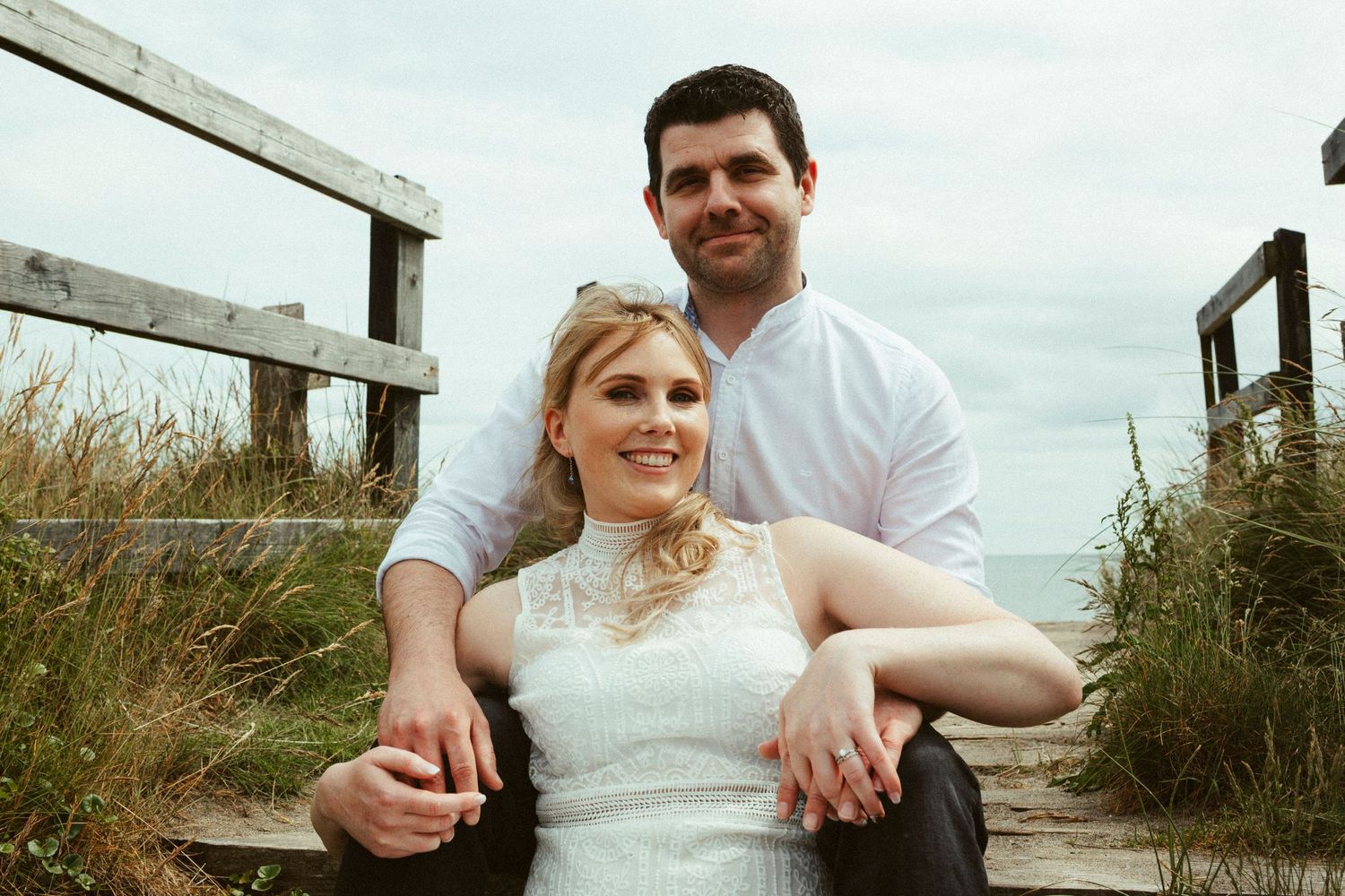 A couple poses together on rustic wooden steps surrounded by tall grasses on their wedding day.