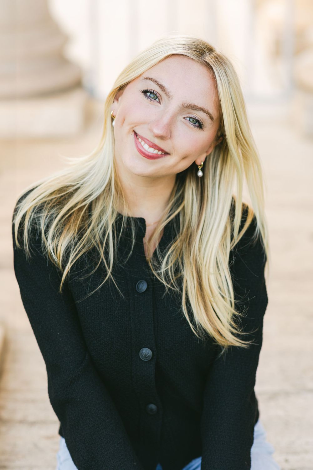 A portrait of someone in a black button-up coat with long blonde hair and a bright smile against a light background.By Chelsee Rawe