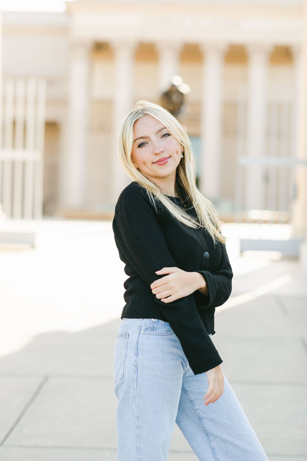 Casual street style outfit featuring light blue jeans and a black sweater on a sunny day with classical architecture behind.By Chelsee Rawe