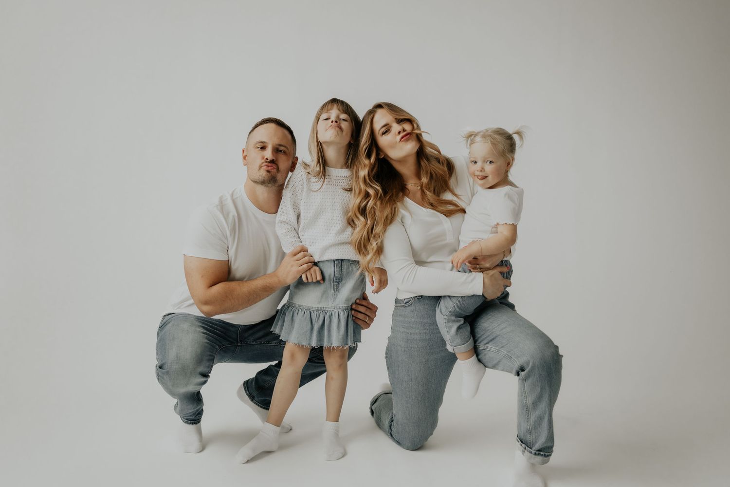 Family with long hair wearing neutral colors sits together on stools against a white studio backdrop.