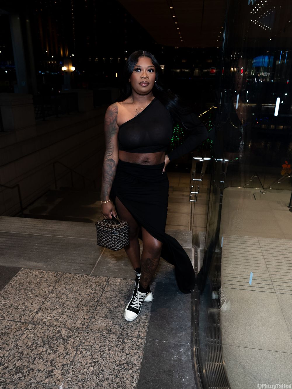 Woman in black crop top and skirt poses against night city backdrop with studded handbag.