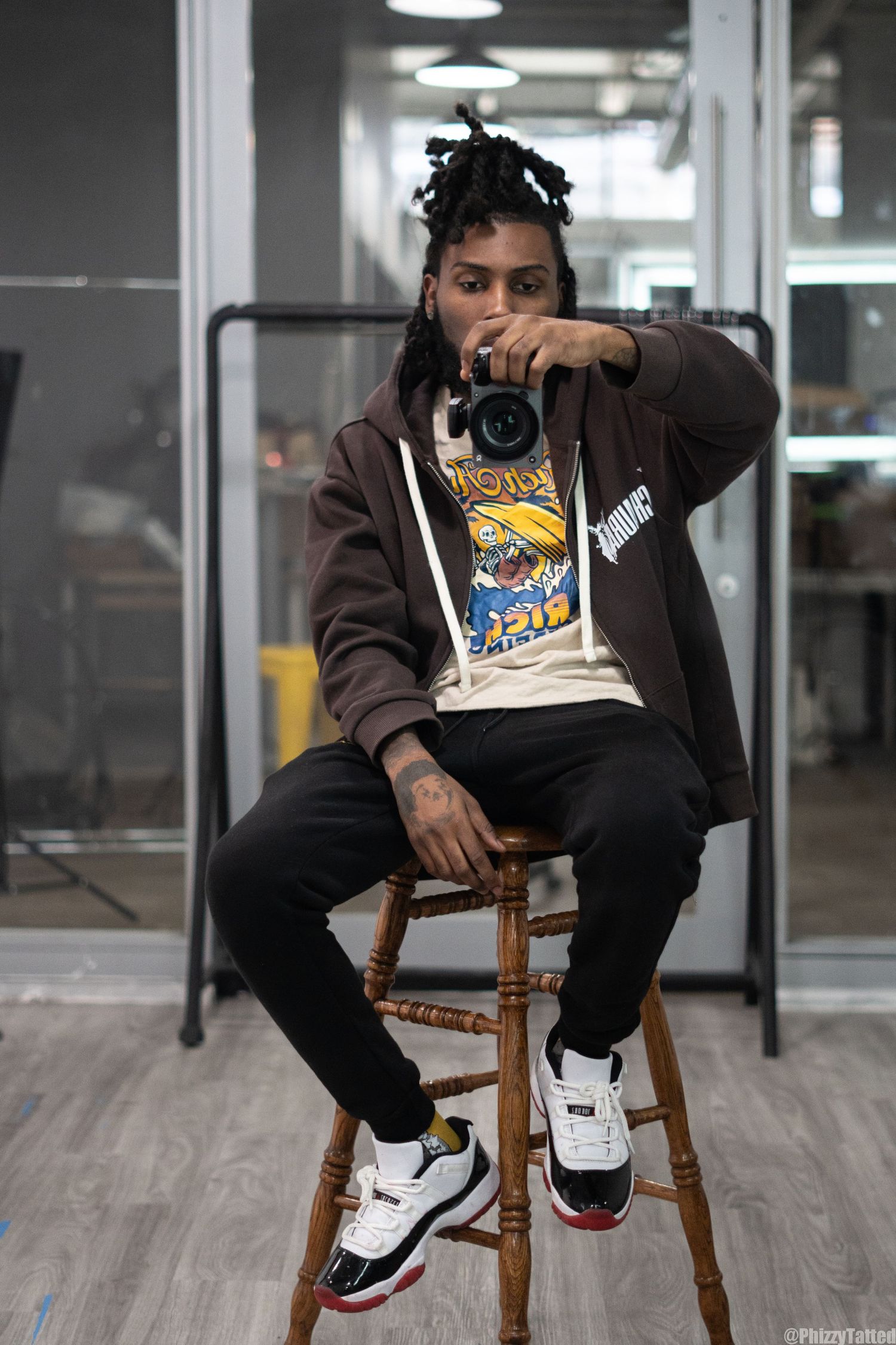 Person sitting on wooden stool wearing dark clothing and sneakers in a studio environment.