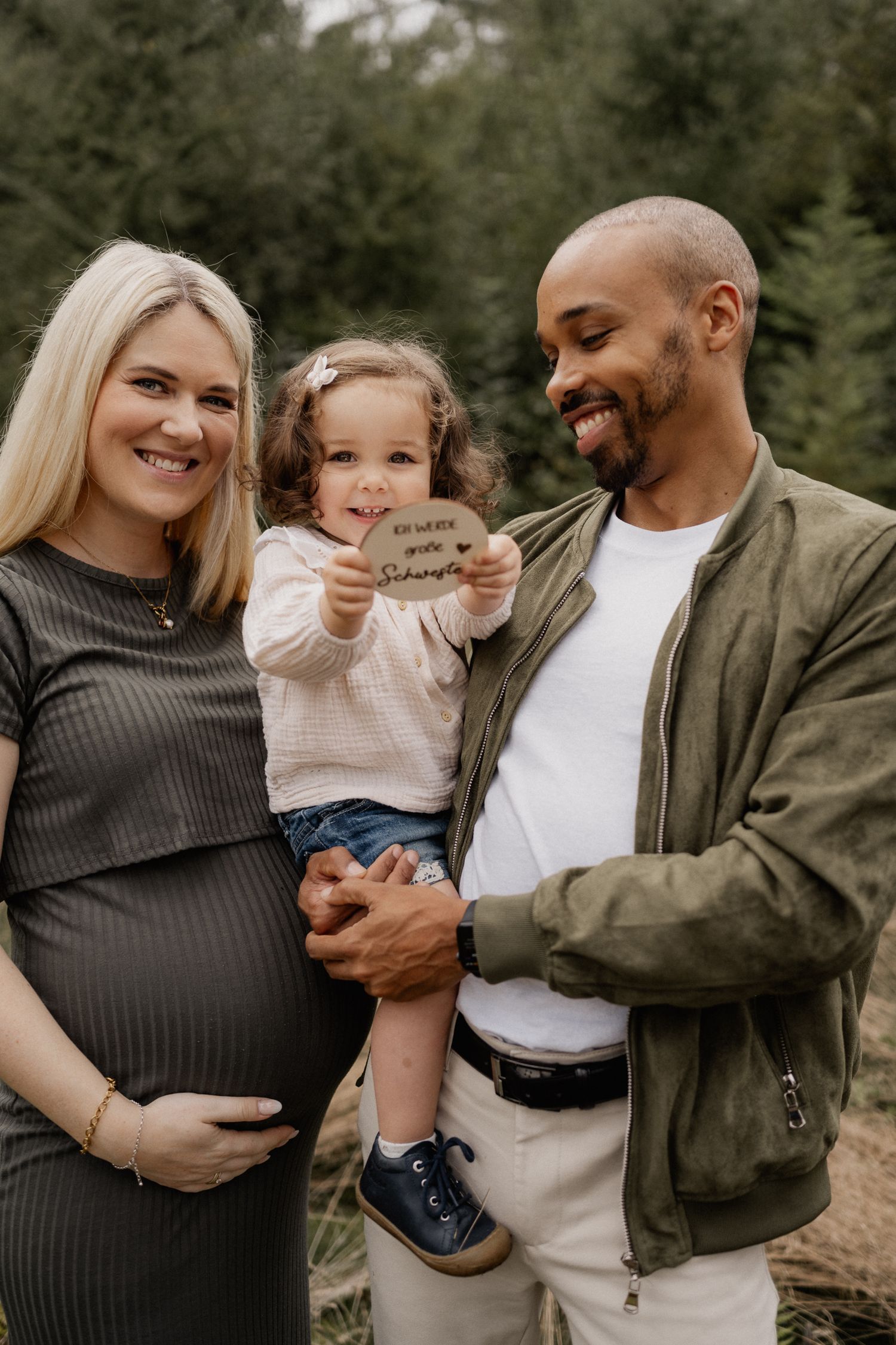 A family poses outdoors with a child holding a small sign while expecting parents embrace in casual fall attire.