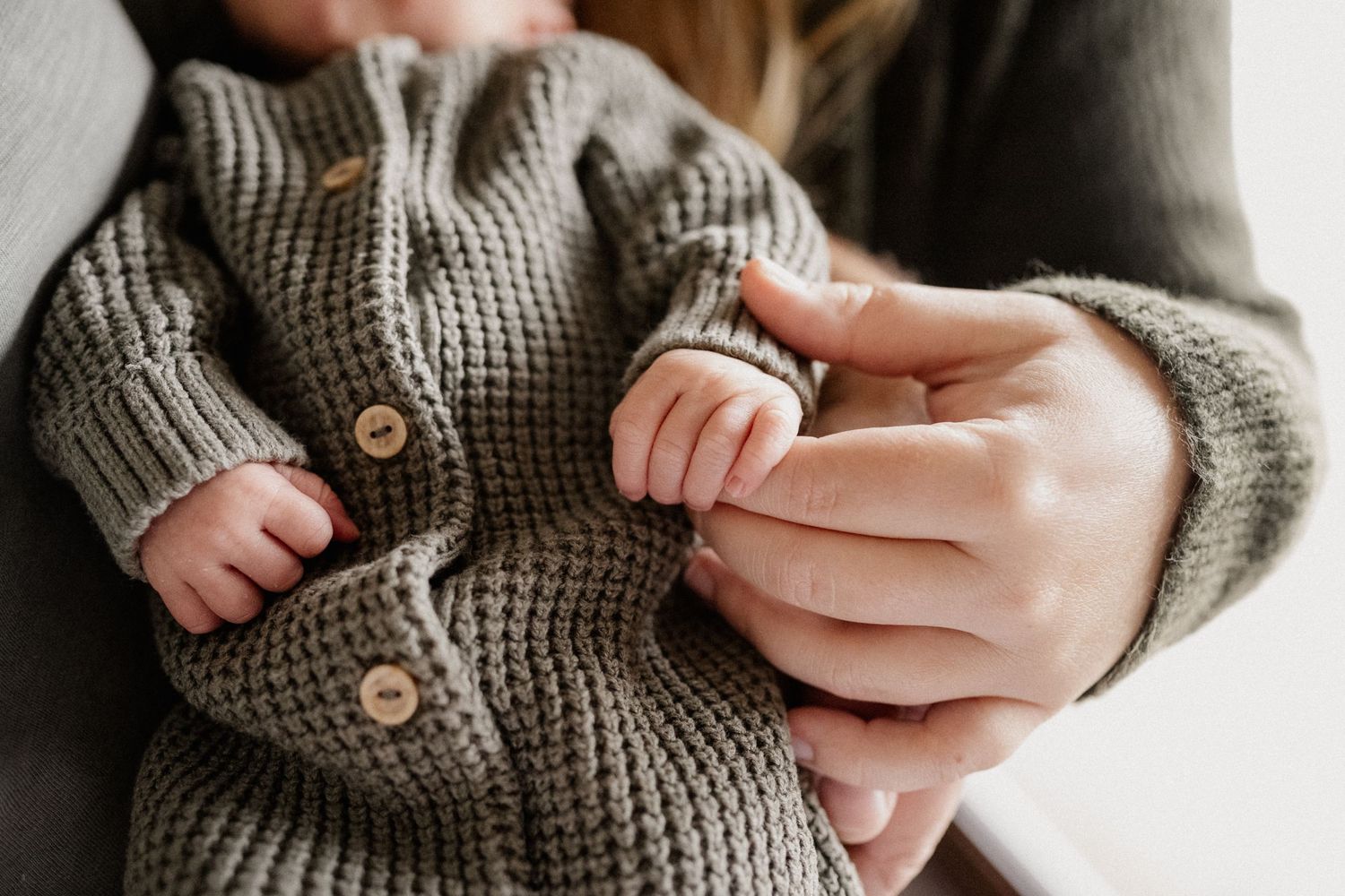 Small hands grasp a cozy grey knit cardigan sweater with wooden buttons in an intimate close-up shot.