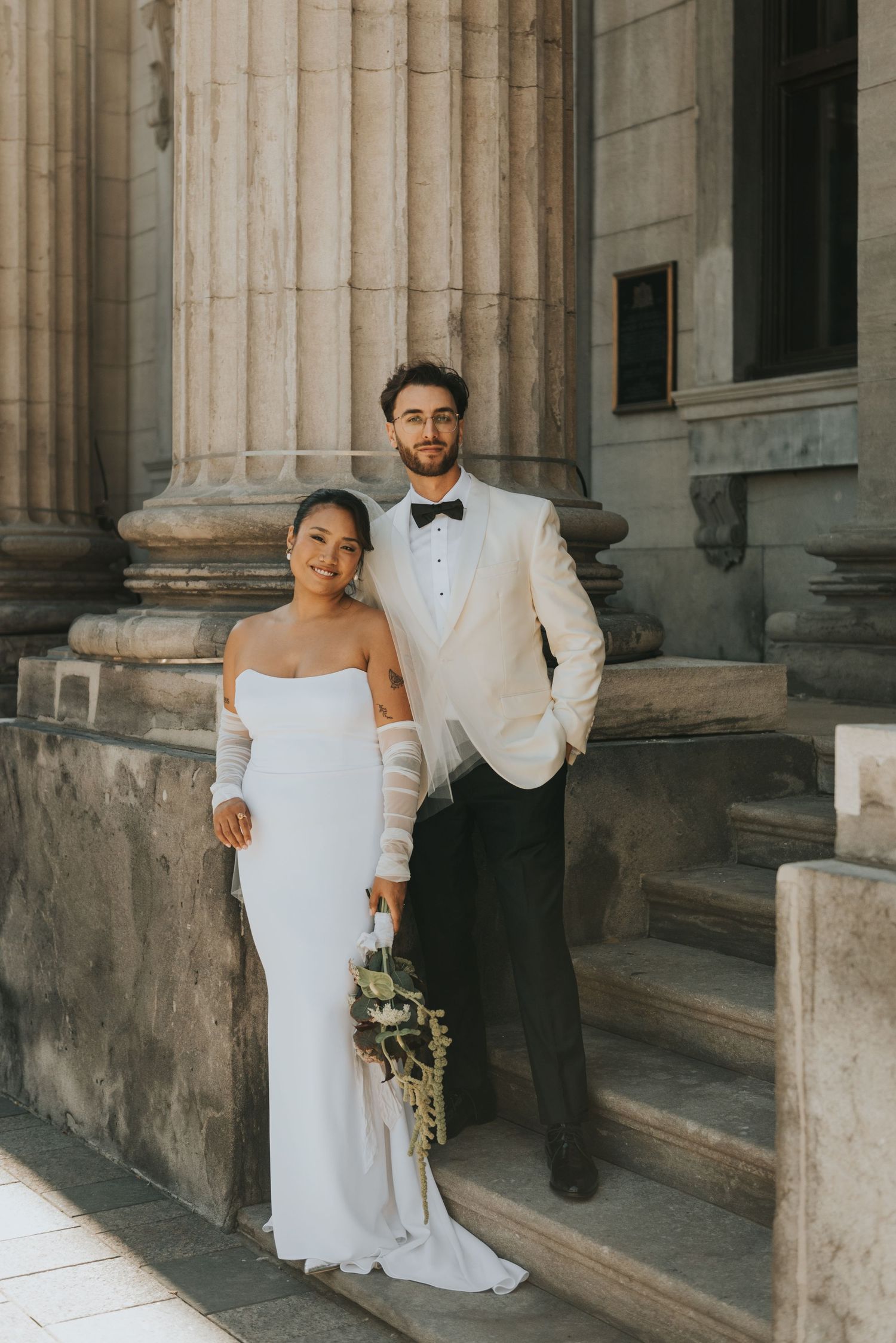 A wedding couple poses elegantly on stone steps beside classical stone columns in warm evening light.