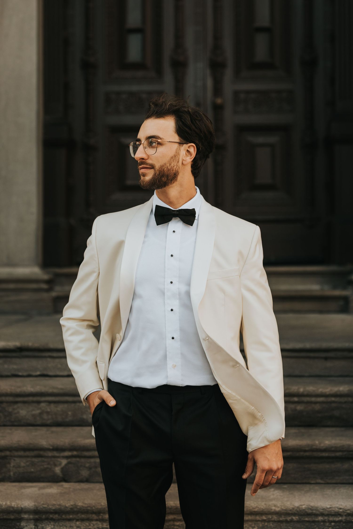 Groom in white dinner jacket and black bow tie stands confidently on stone steps with architectural backdrop.