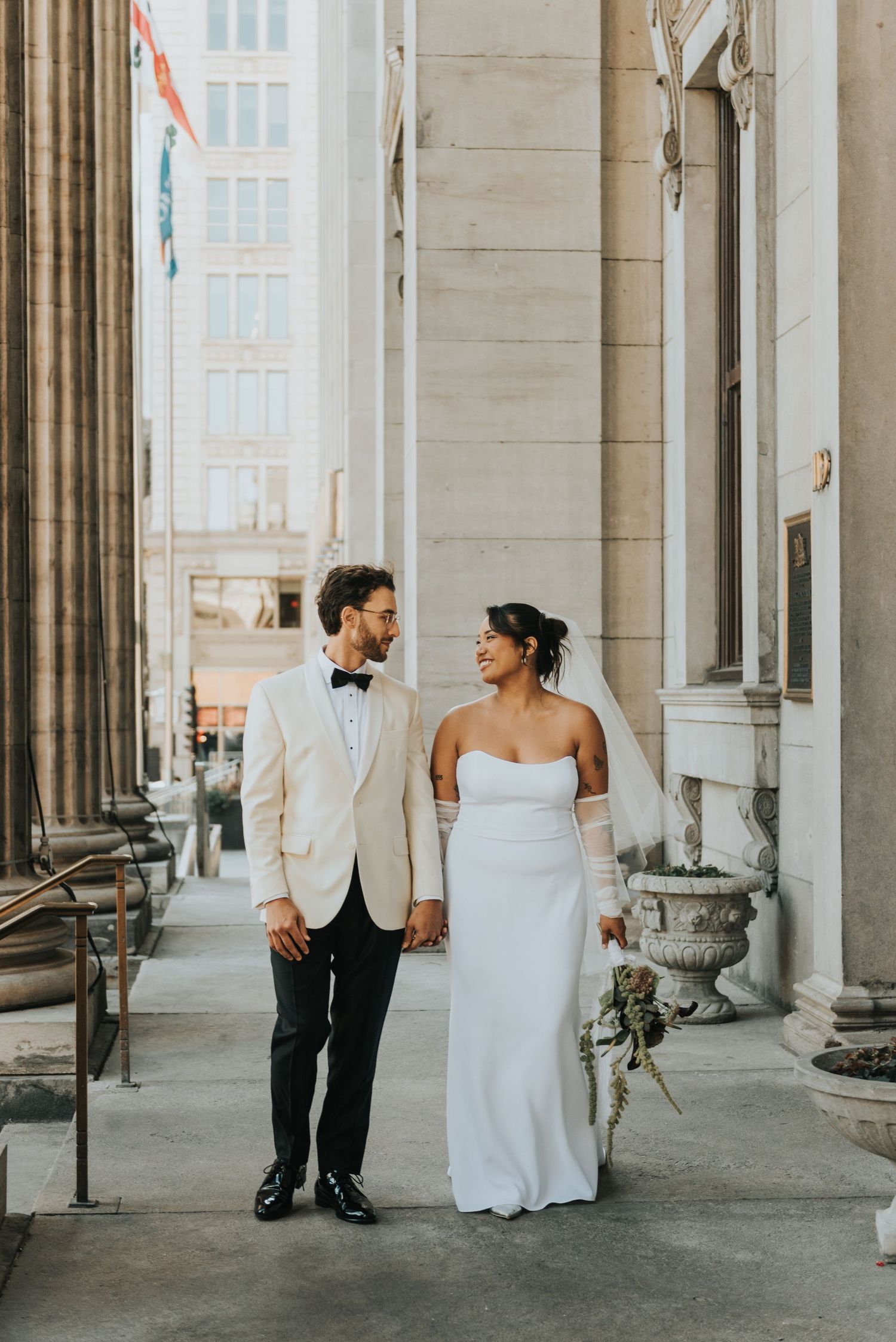 A couple dressed in wedding attire holds hands while standing between elegant stone columns of a historic building.