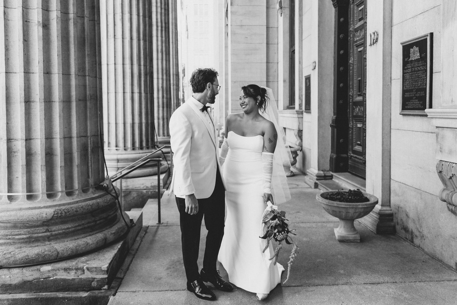 A black and white wedding photo of a couple standing between classic stone columns of an elegant building.