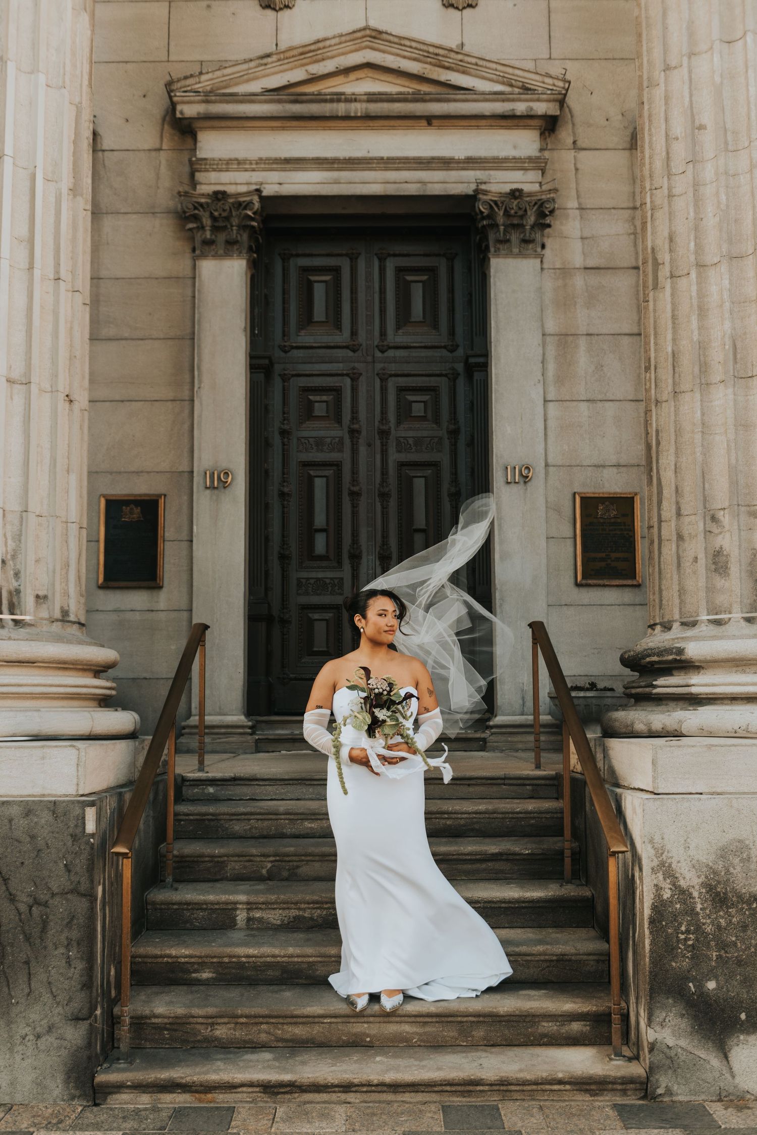 A bride in an off-shoulder white dress holds a bouquet while standing on stone steps of a grand historic building entrance.