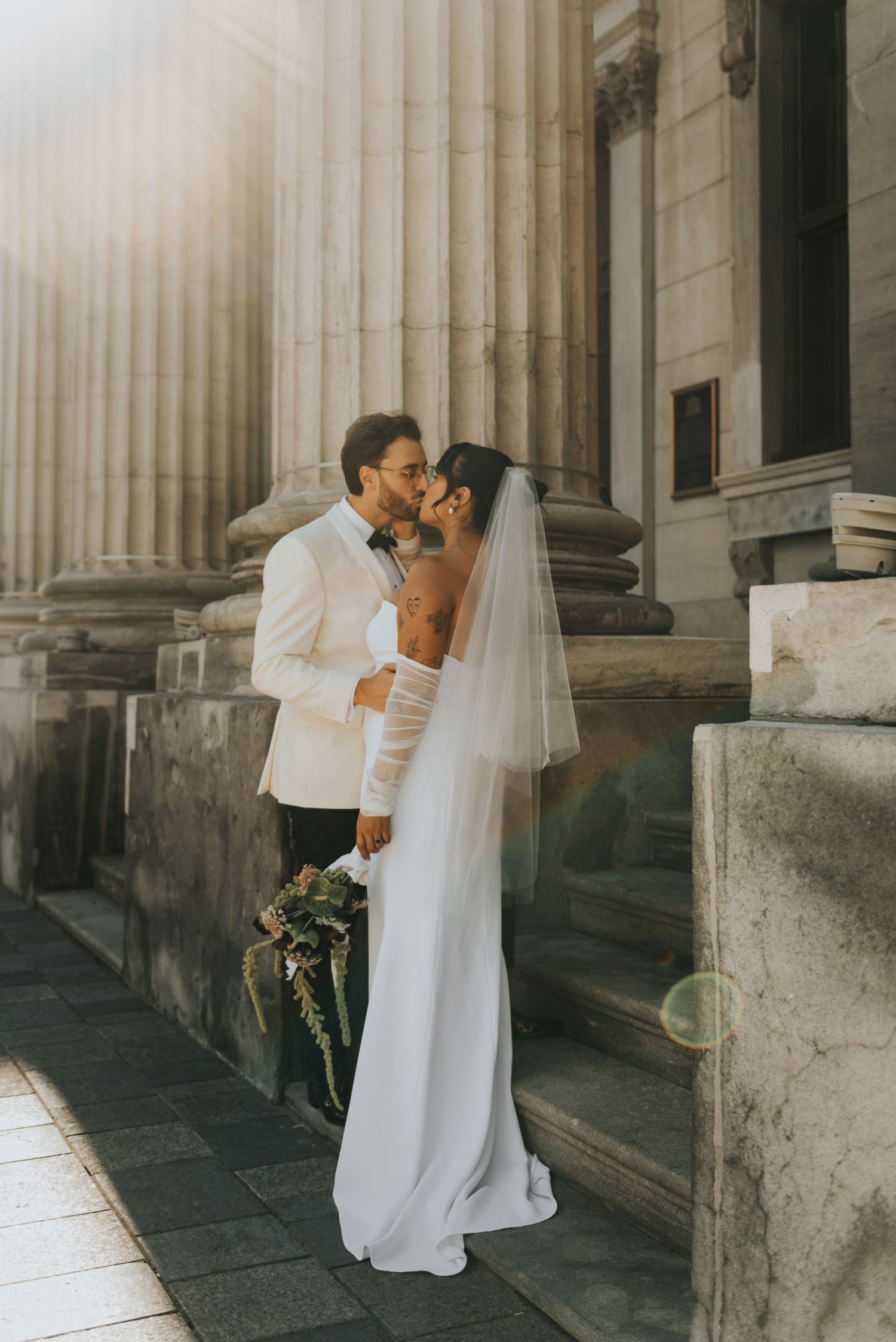 A couple shares an intimate moment in front of stone columns on their wedding day, with bride in white gown and flowing veil.