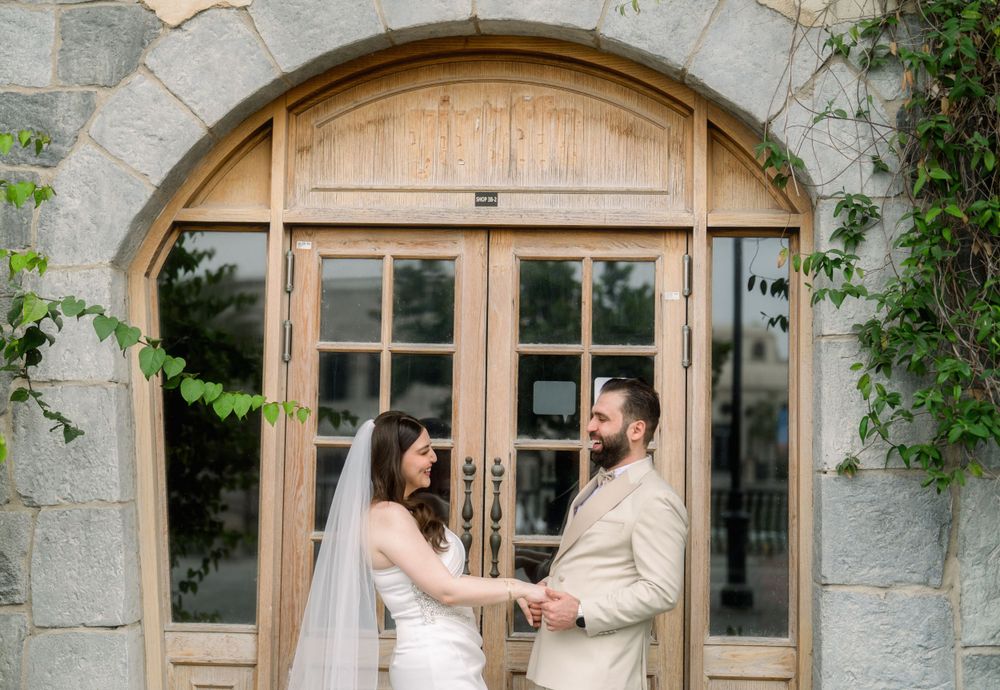 A couple in light-colored outfits holds hands in front of an arched wooden doorway with stone walls and climbing vines.