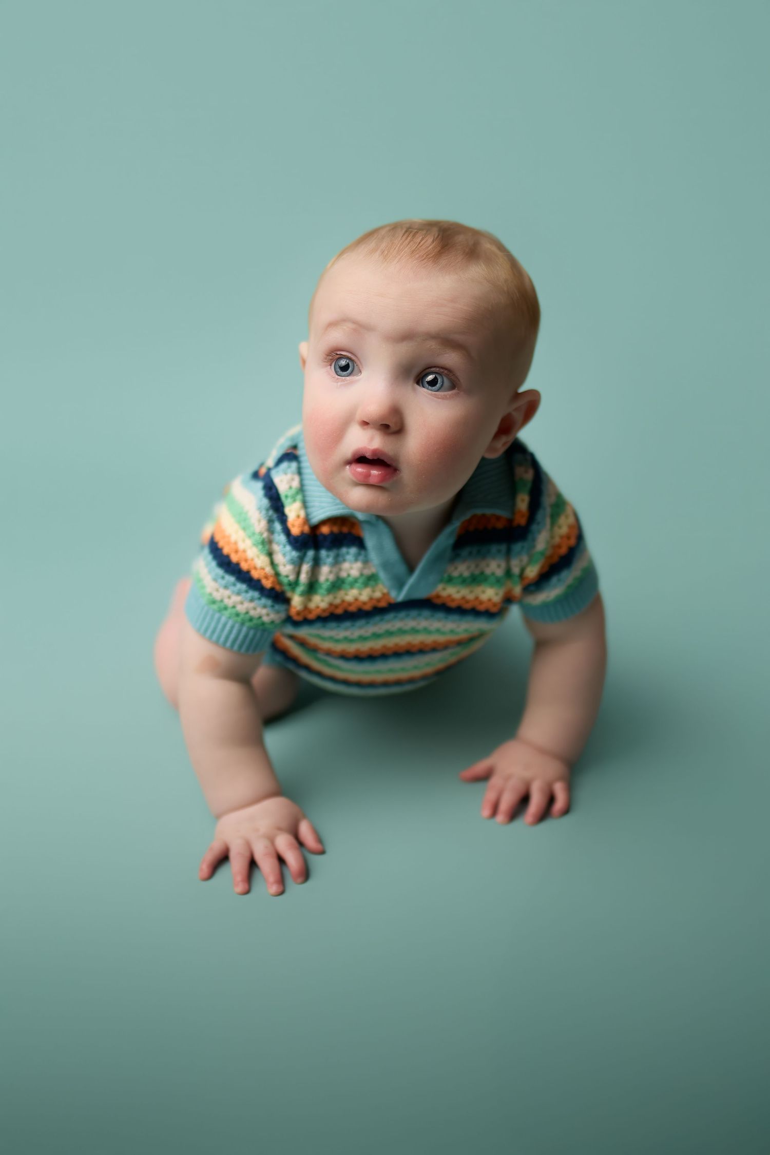 A baby in a striped colorful sweater crawls on a teal backdrop during a studio photoshoot.