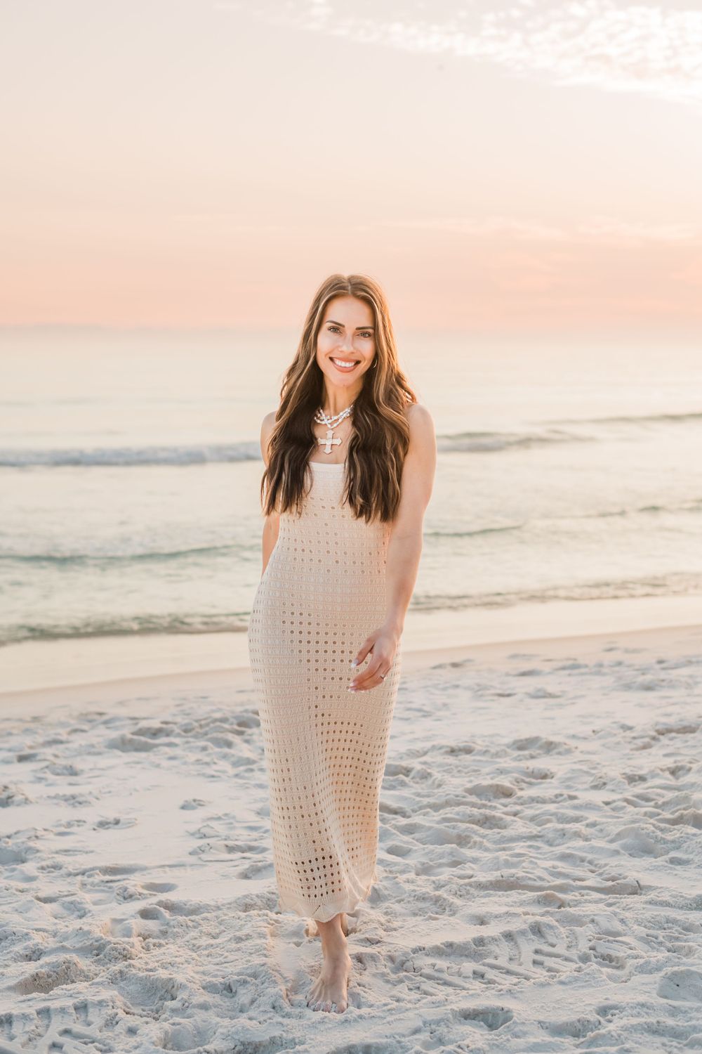 Woman in crochet maxi dress smiles while standing barefoot on sandy beach at sunset.