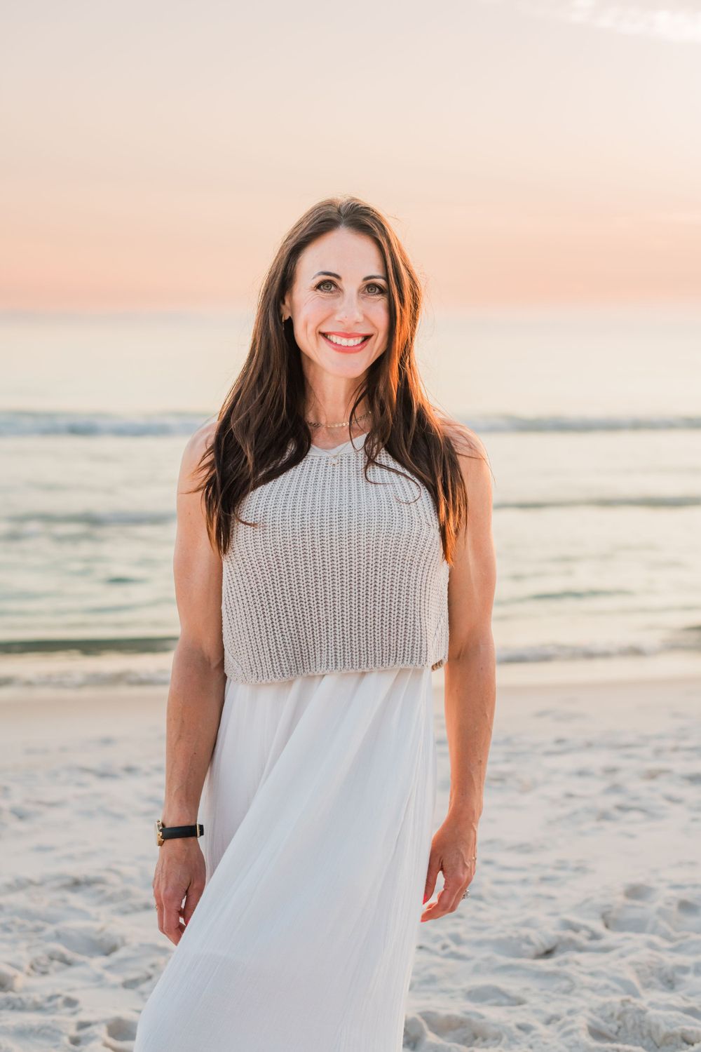 Person in a knitted crop top and white dress poses on a sandy beach at sunset with ocean waves in the background.