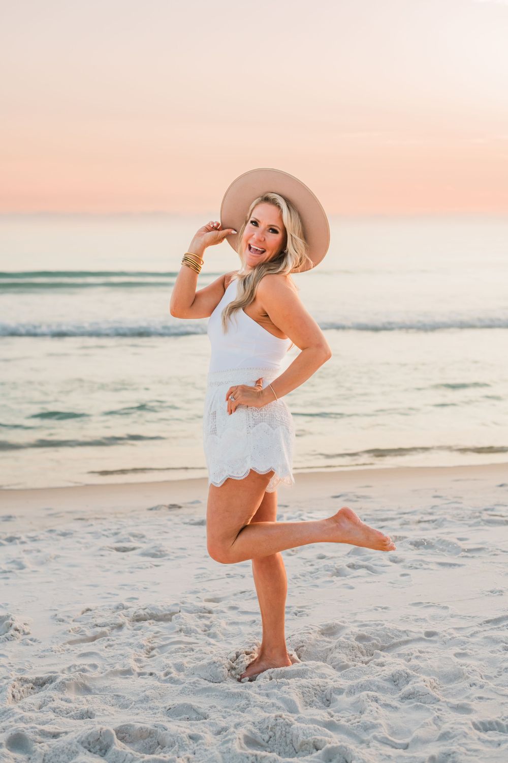 Person in a white summer dress and floppy hat poses playfully on a beach at sunset with pink sky and ocean waves.