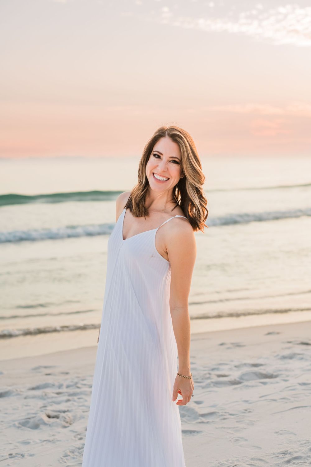 A beachgoer in a flowing white sundress smiles while standing on sandy shores at sunset.