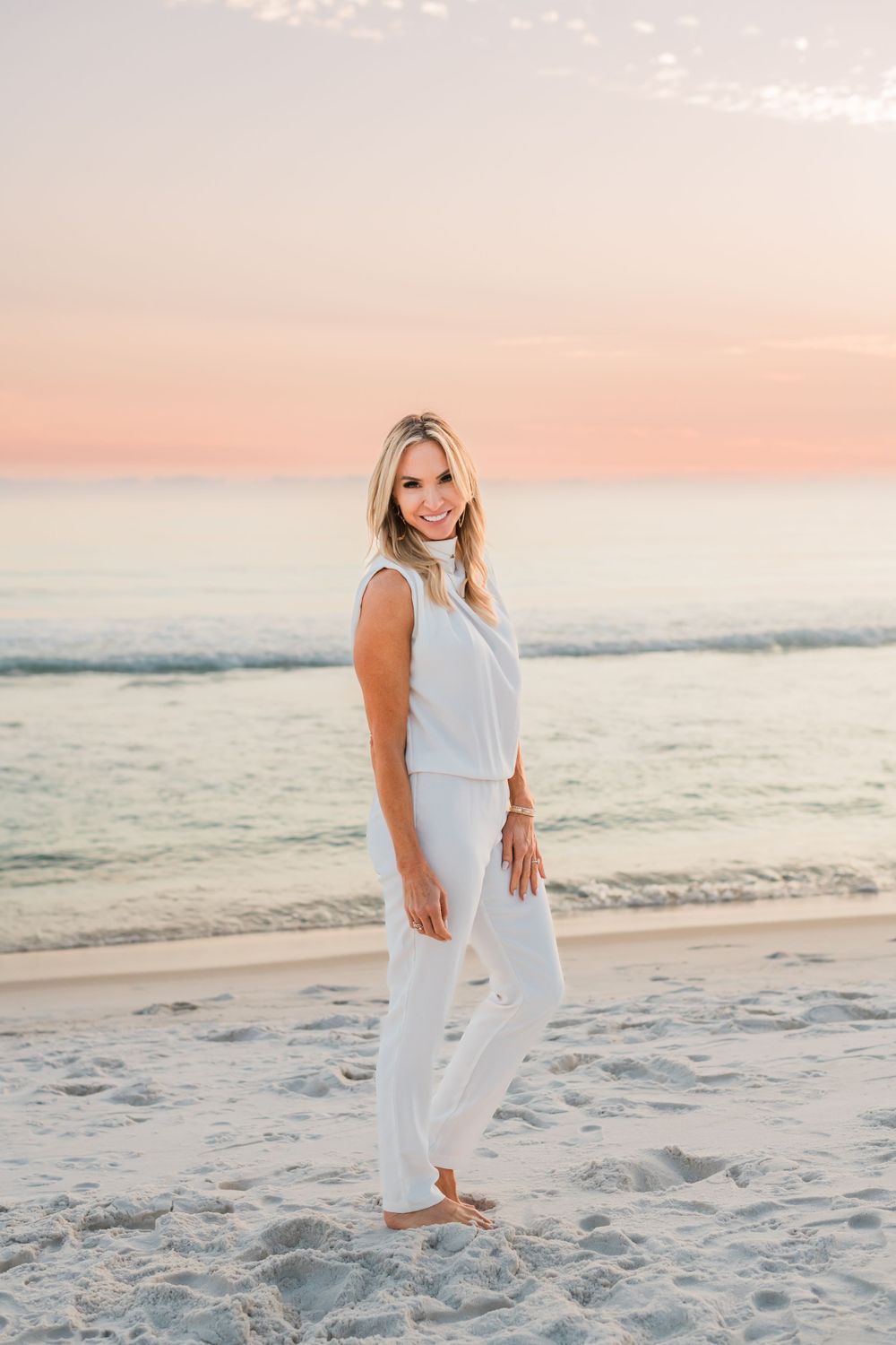 A person in an all-white outfit poses on a sandy beach at sunset with a pink and orange sky.