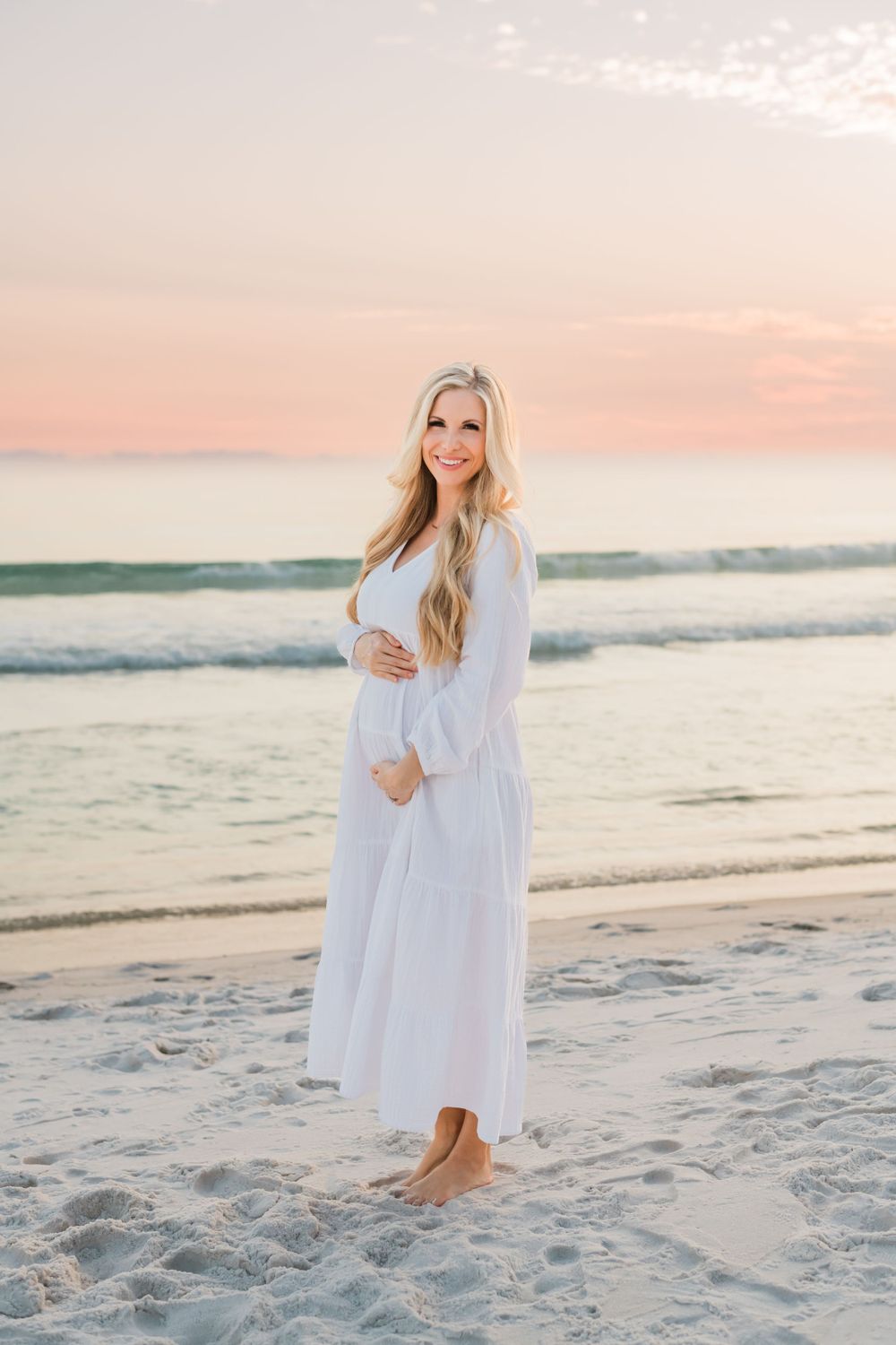 A woman in a flowing white dress stands barefoot on a sandy beach at sunset with gentle waves in the background.
