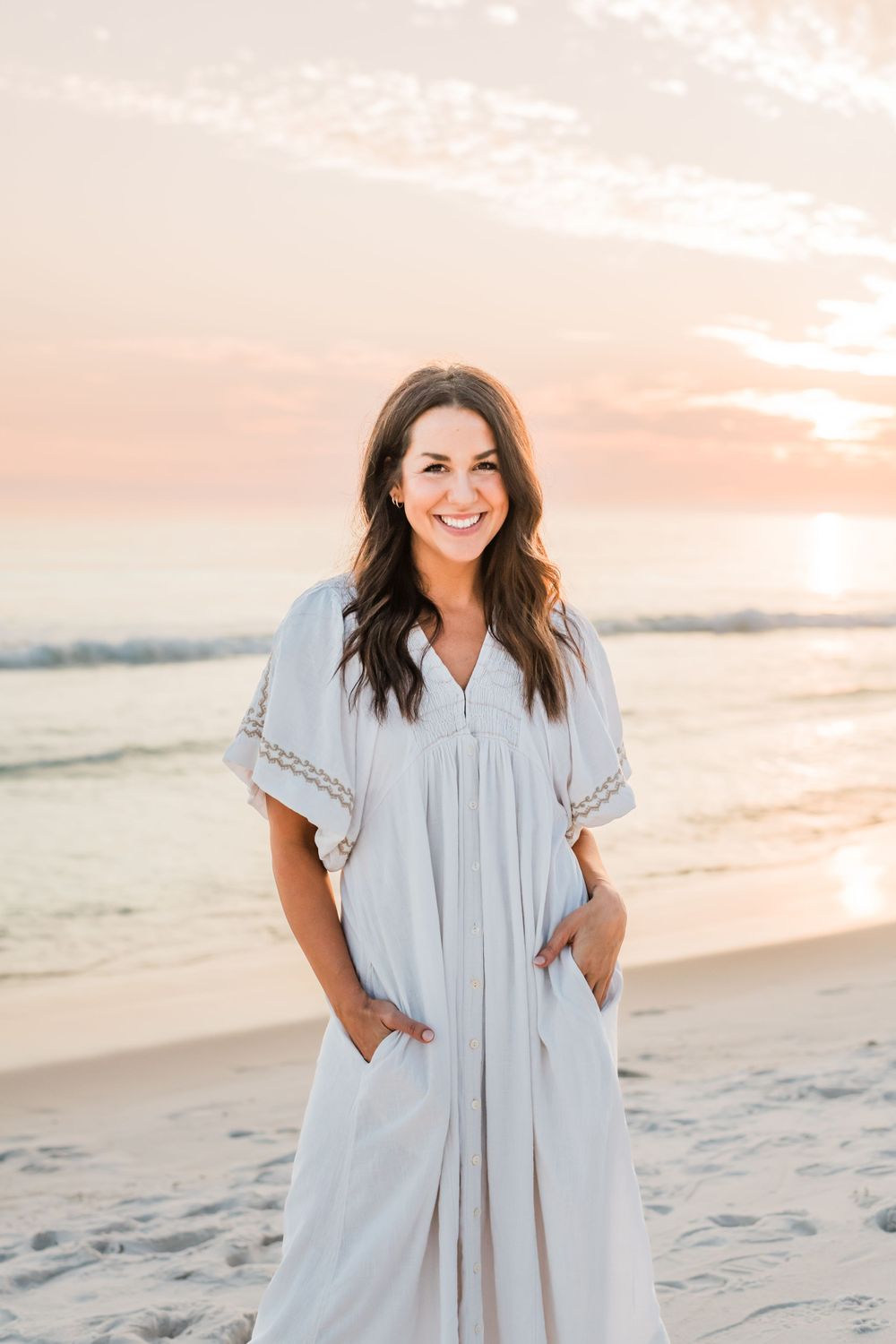 Woman in flowy white dress smiles while standing on beach at sunset with pink sky and ocean waves behind her.