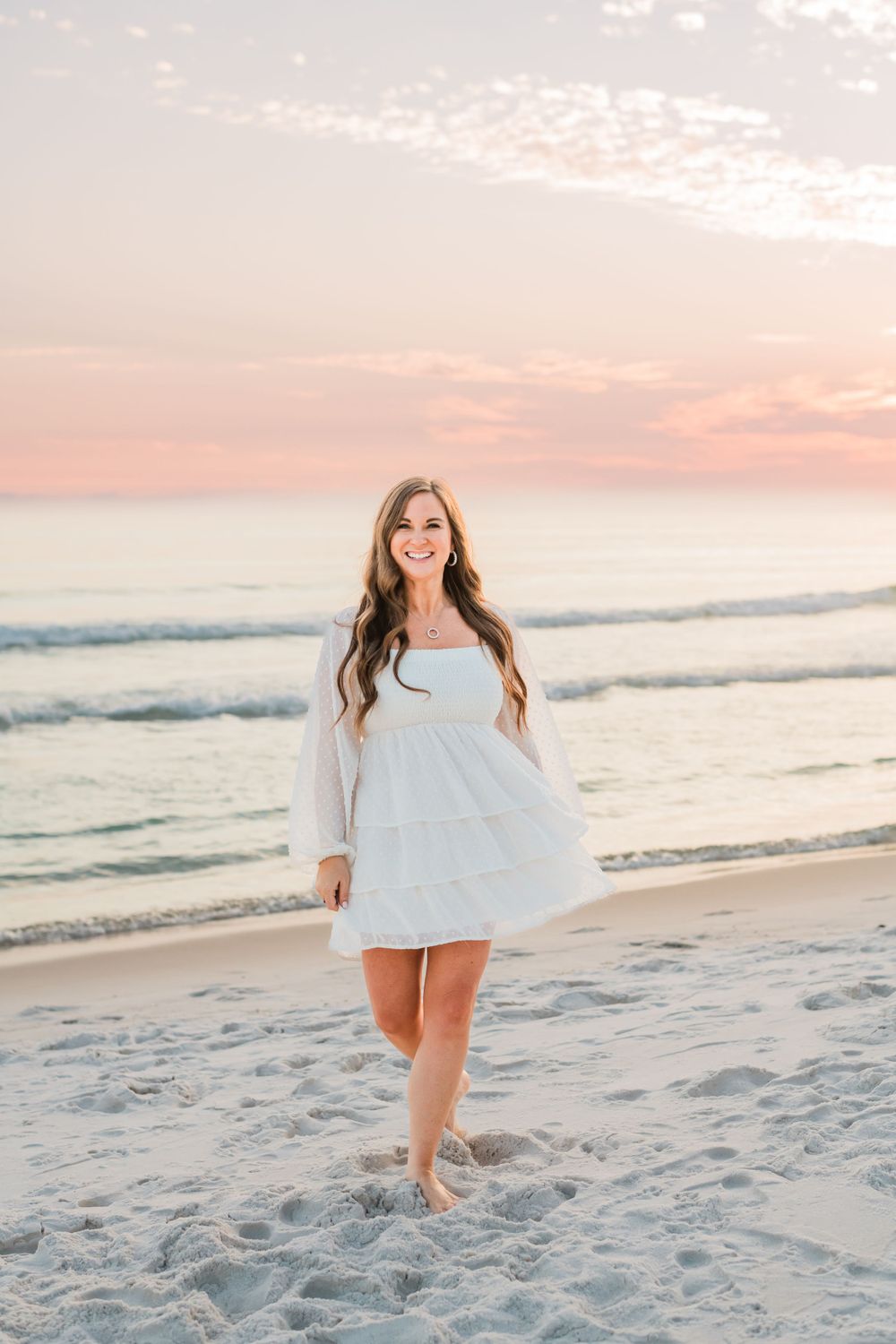 Woman in flowing white dress walks barefoot on beach at sunset with pink sky and ocean waves in background.
