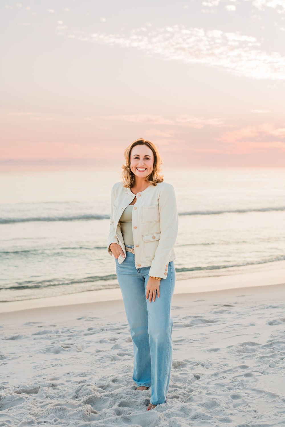 A person in a cream blazer and light jeans stands on a beach at sunset with pink sky and ocean waves in the background.