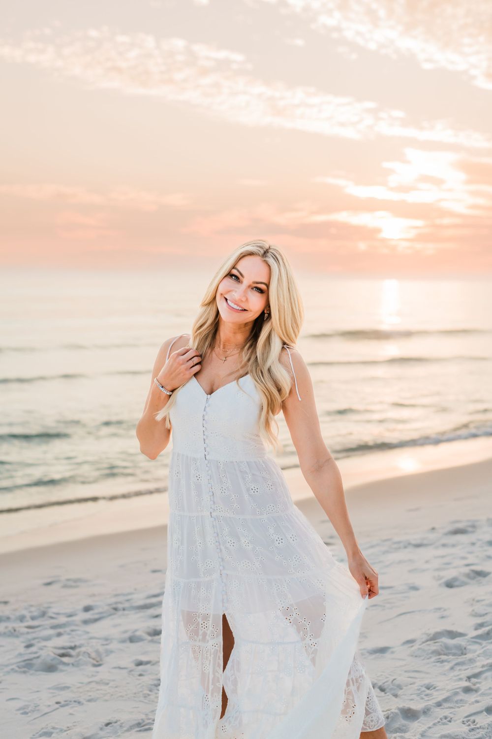 A person wearing a white lace maxi dress poses on a sandy beach at sunset with waves in the background.