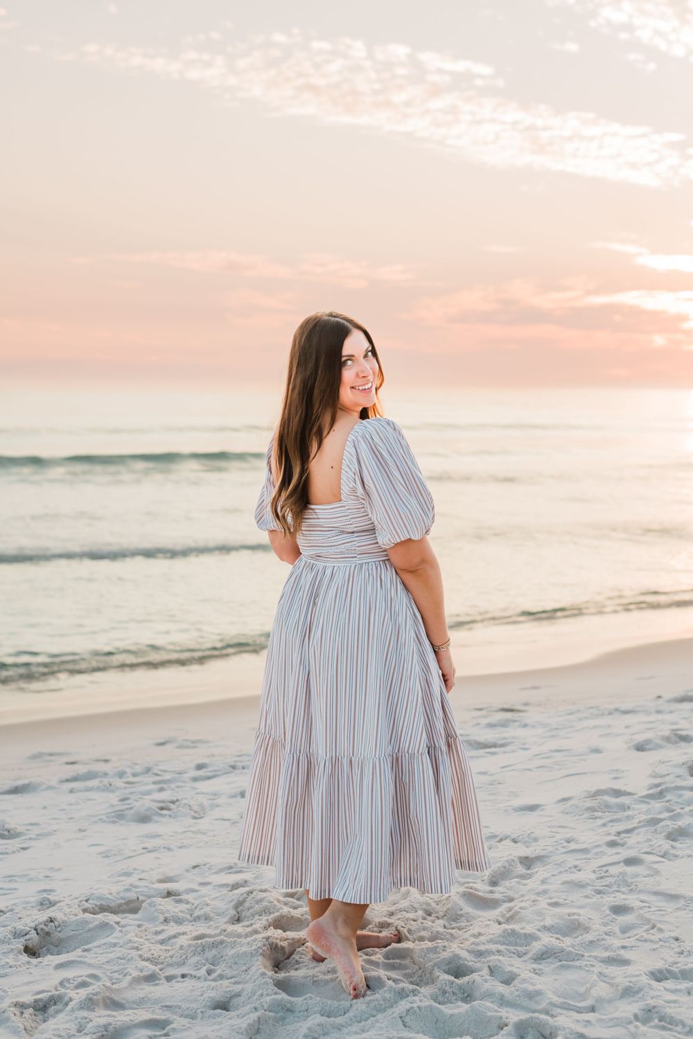 Woman in striped summer dress poses on sandy beach at sunset with ocean waves in background.