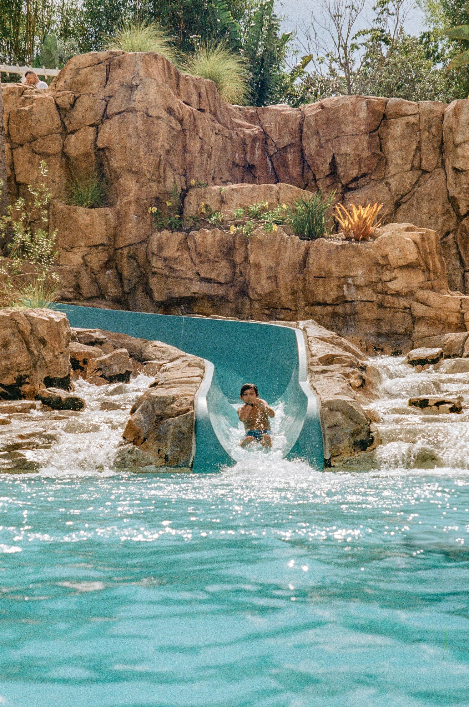 A turquoise water slide curves through artificial rock formations with cascading waterfalls in a tropical pool setting.