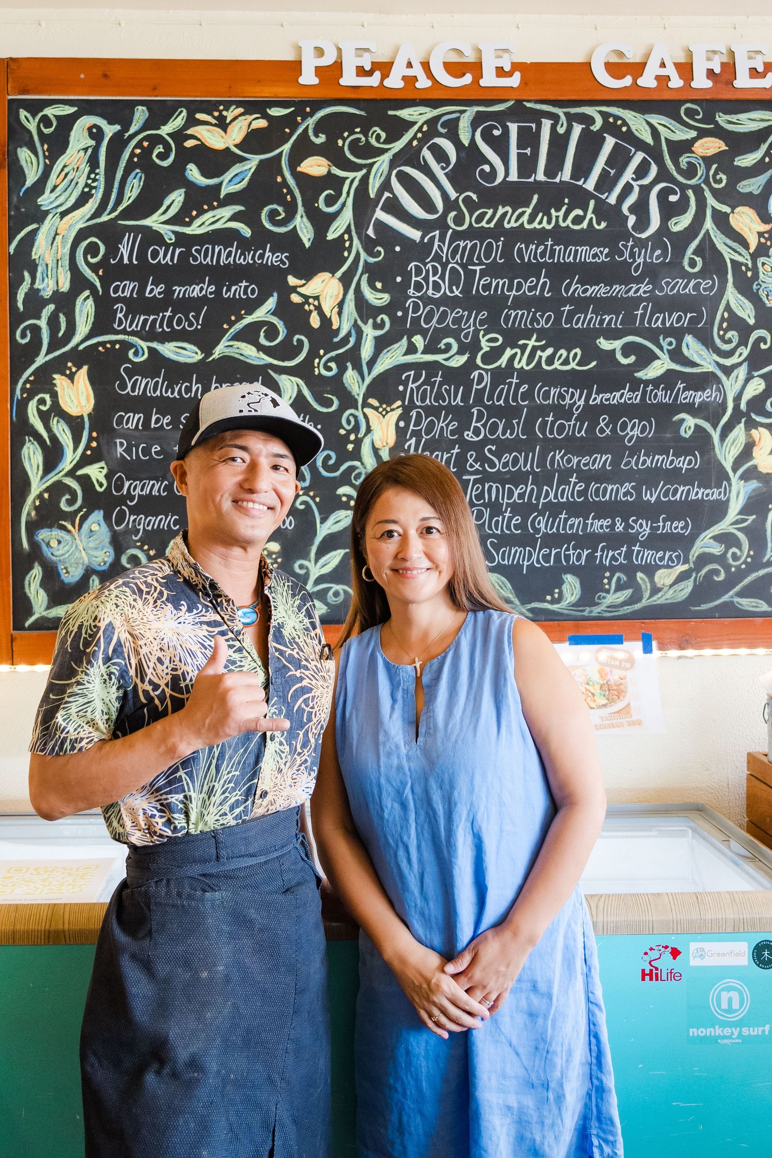 Two restaurant staff members stand in front of a chalkboard menu at Peace Cafe with decorative floral designs.