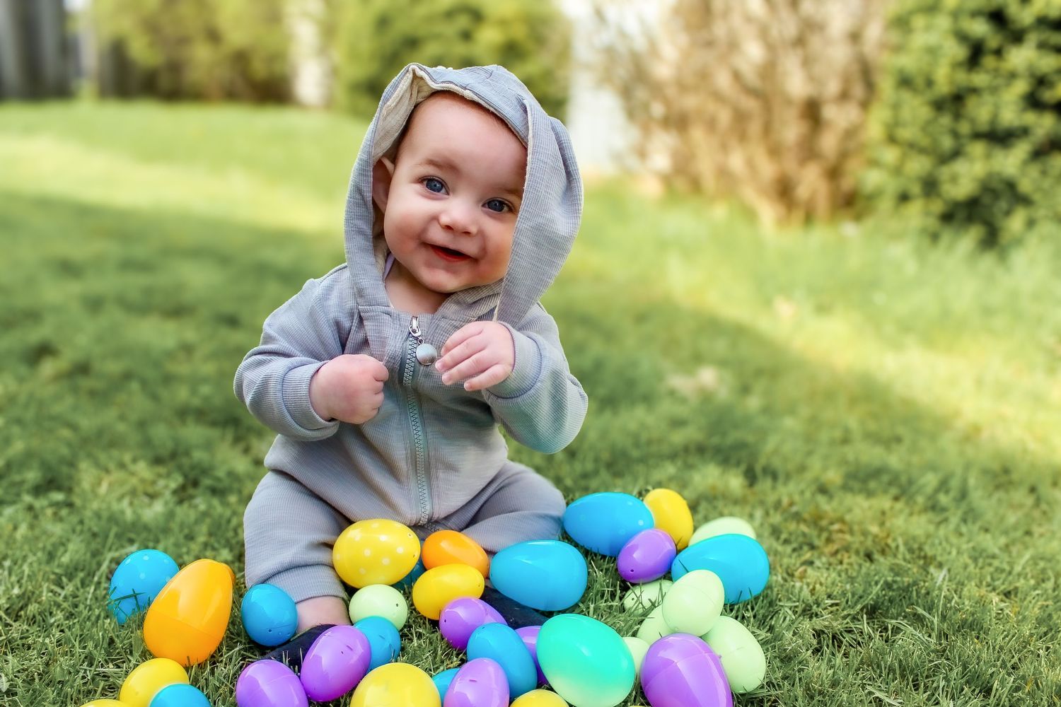 Springfield, IL Photographer captures a baby sitting in the grass with Easter eggs while wearing a bunny onesie and smiling.