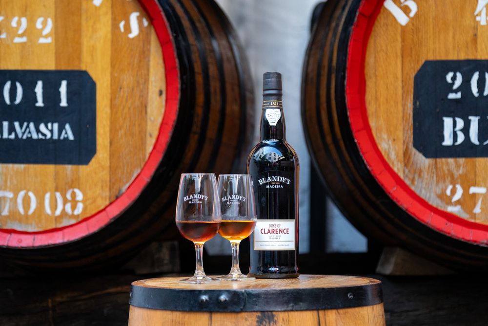Bottle of port wine and two glasses displayed on wooden barrel with vintage wine casks in background.