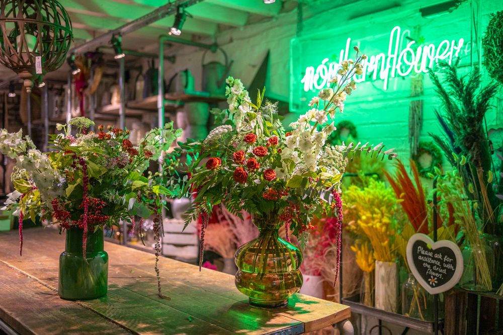 Glass vases filled with colorful dried flowers and greenery displayed under a green neon sign in a rustic setting.