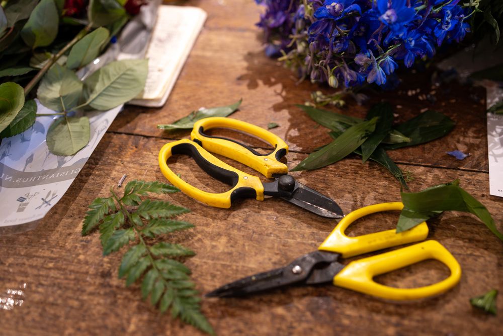 Yellow gardening scissors with bright blue flowers and green foliage on rustic wooden surface.