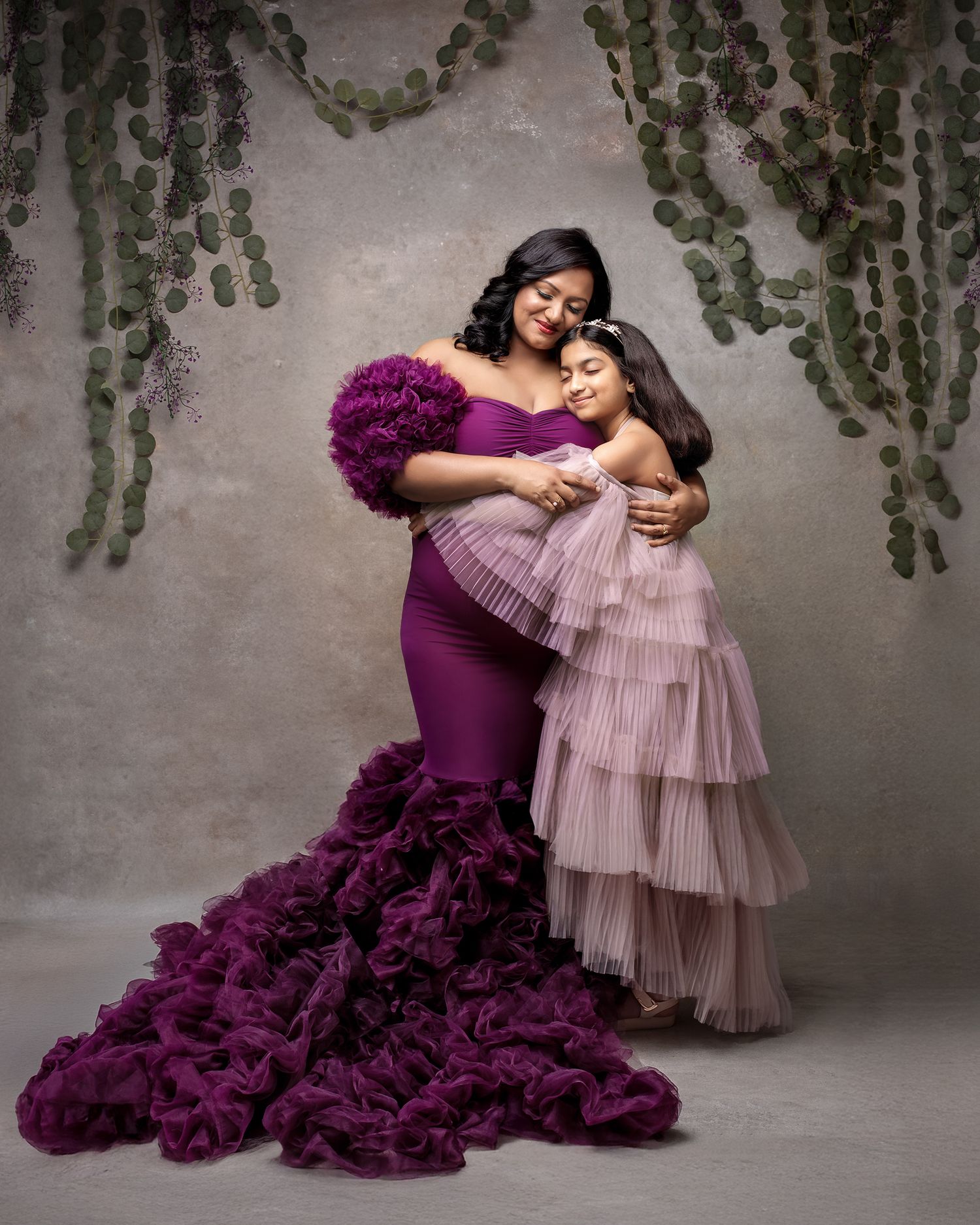Motherhood celebrated in style: mum in a purple gown, daughter in a pleated mauve couture gown, embracing in a fine art studio portrait.