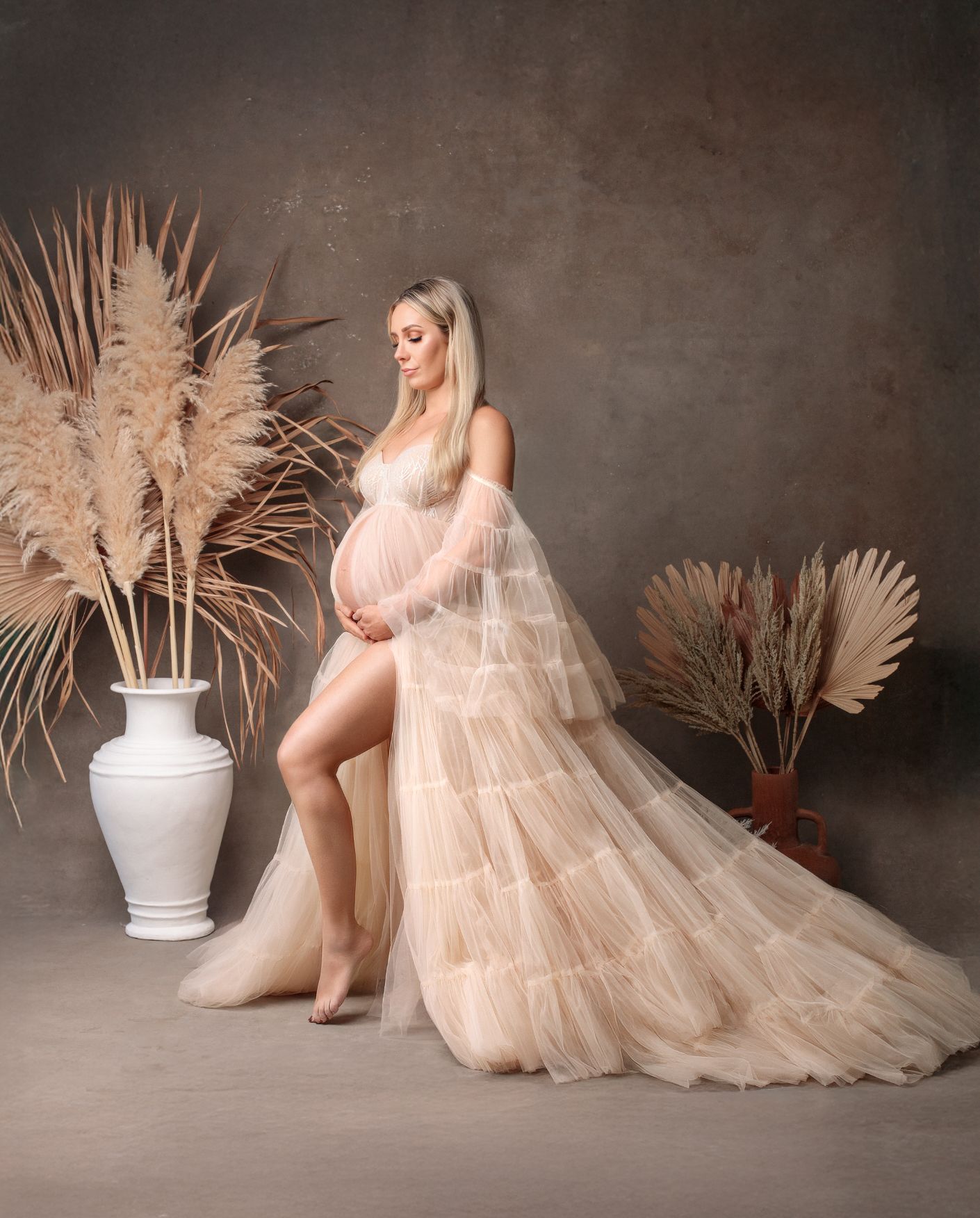 A beautiful blonde holds her pregnant belly wearing a striking gown with a long train and sparkly bust detail against brown backdrop.