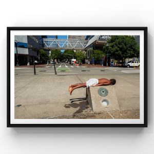 Someone performs a creative plank pose across a concrete bench in a sunny urban plaza with buildings and trees in background.