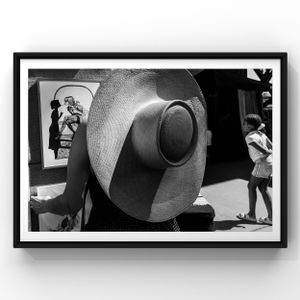 A vintage black and white street photograph featuring a large straw hat in the foreground and silhouettes in background.