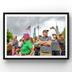 Group of spectators gather outdoors with American flag and skyscrapers visible in background.