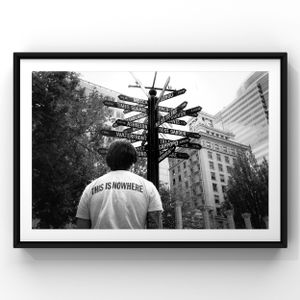 Person wearing 'THIS IS NOWHERE' shirt looks up at directional street sign post in black and white.